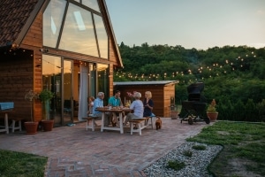 Group of adults and a child sharing a meal at a wooden house patio at sunset, string lights overhead.