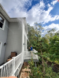 Person using a hose to spray water against the side of a gray house, standing on a deck with white railing, trees and blue sky in the background.