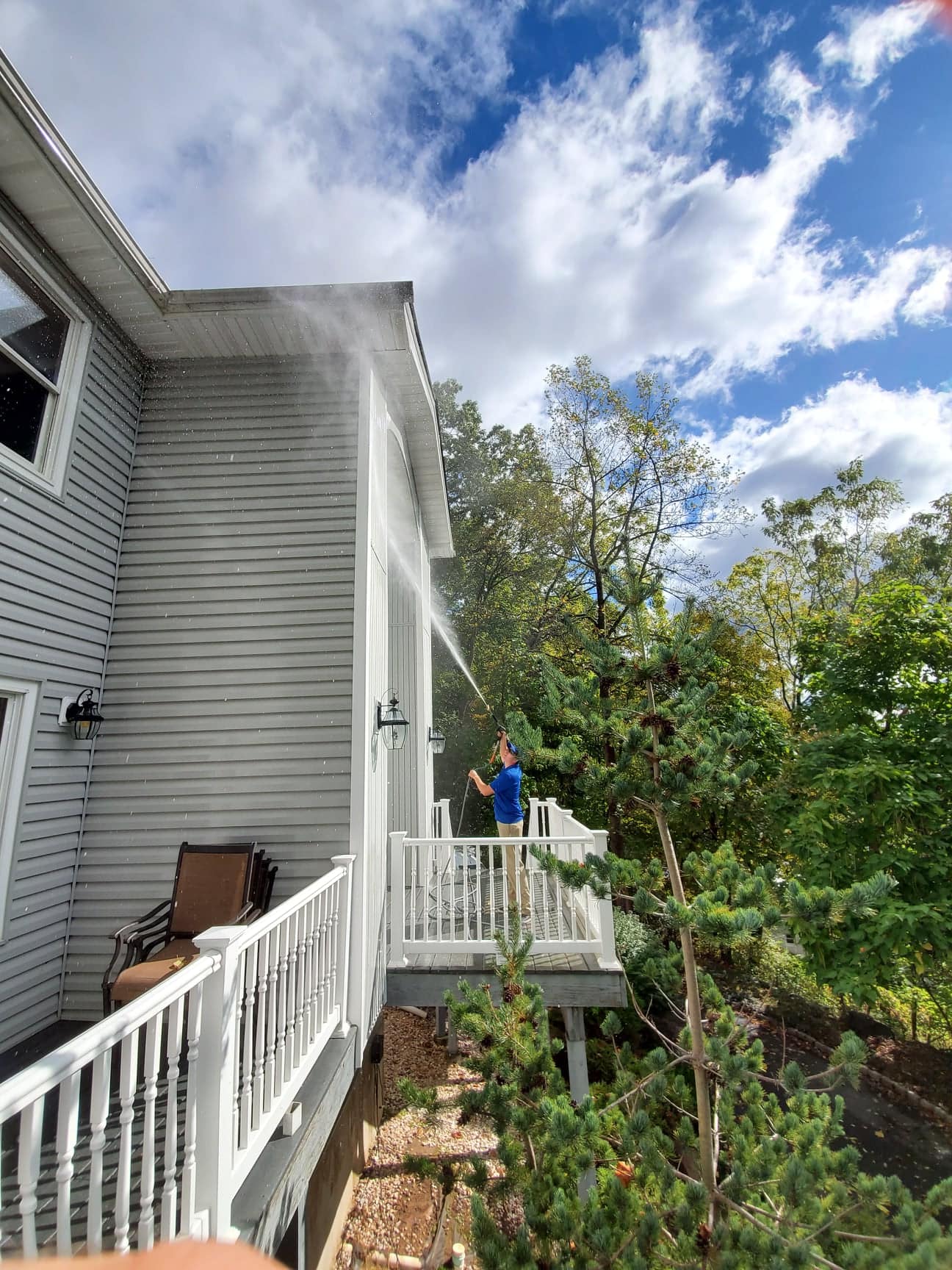 Person using a hose to spray water against the side of a gray house, standing on a deck with white railing, trees and blue sky in the background.